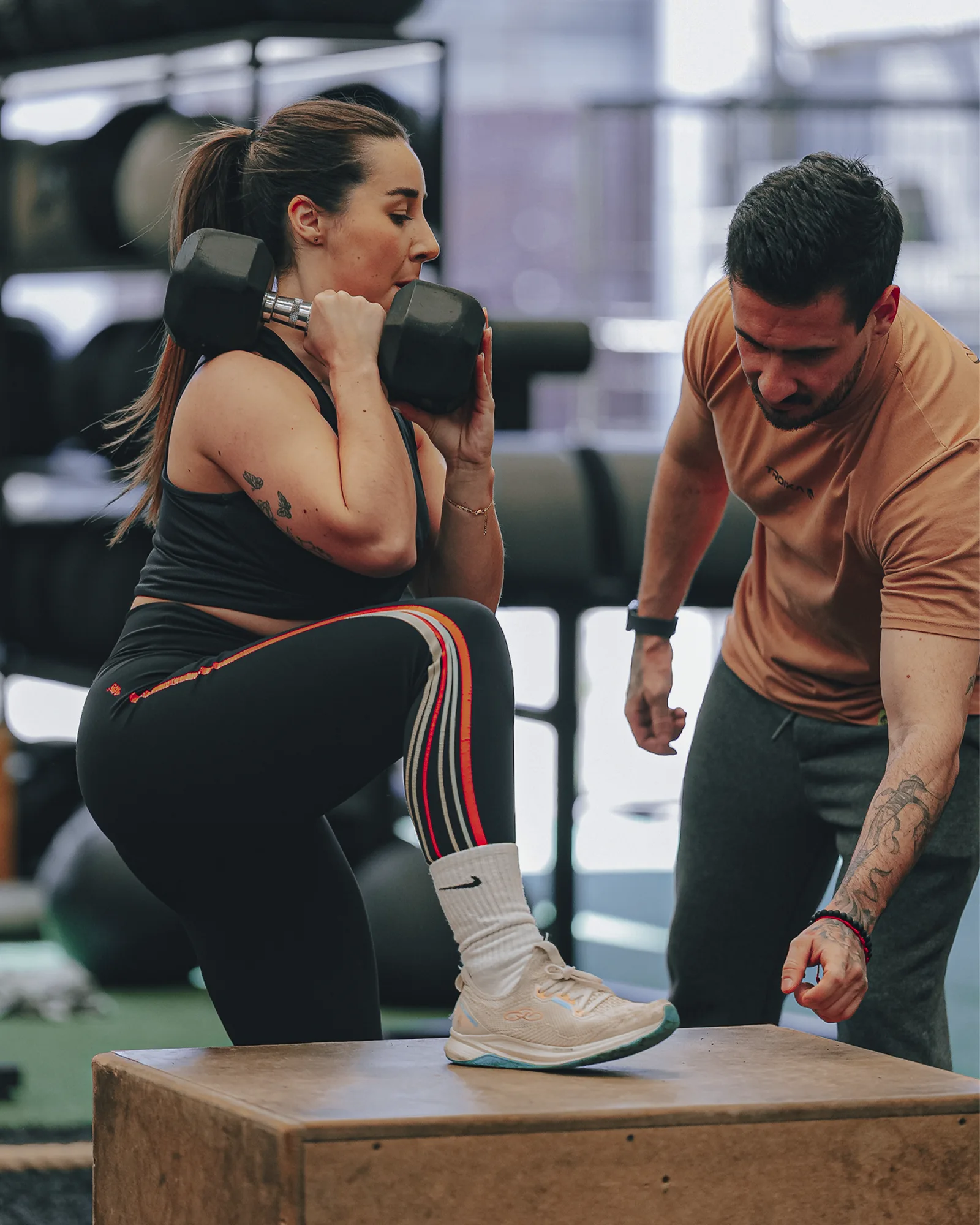 Coach orientando uma aluna durante um exercício com halteres e subida no caixote